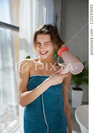 Smiling young woman wearing tower, posing, while drying wet hair beside window. Front view of beautiful girl with haircut winking to camera in bathroom in sunny morning. Lifestyle, routine concept. Smiling young woman wearing tower, posing, while drying wet hair beside window. Front view of beautiful girl with haircut winking to camera in bathroom in sunny morning. Lifestyle, routine concept. 110293085