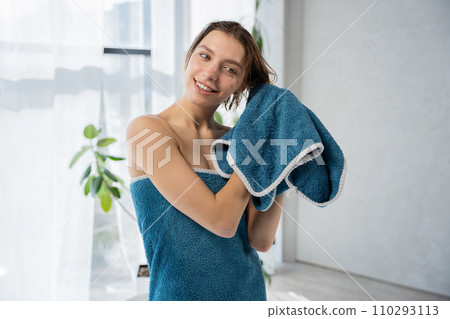 Attractive blonde female drying short, wet hair with towel in bathroom at home. Front view of smiling caucasian woman caring hair after shower against sunlit window. Concept of beauty care, lifestyle. Attractive blonde female drying short, wet hair with towel in bathroom at home. Front view of smiling caucasian woman caring hair after shower against sunlit window. Concept of beauty care, lifestyle. 110293113