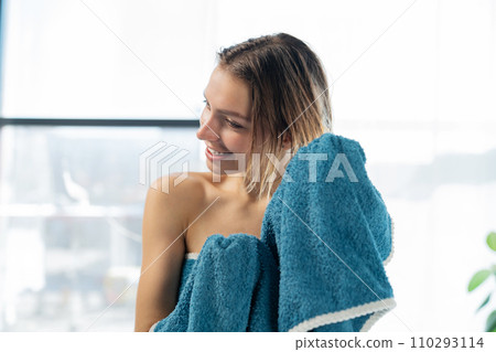 Happy young female drying hair with turquoise cotton towel after showering against wide window. Portrait of smiling caucasian woman using towel on wet head in bathroom. Concept of hair care, beauty. Happy young female drying hair with turquoise cotton towel after showering against wide window. Portrait of smiling caucasian woman using towel on wet head in bathroom. Concept of hair care, beauty. 110293114