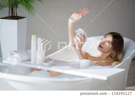 Pretty caucasian woman playing with sun beam, while bathing at home. Side view of joyful blonde girl washing arms, while laying in comfortable bathtub, in front of grey wall. Body care concept.  110293127