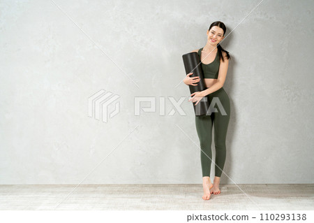 Beautiful brunette sportswoman posing, while carrying yoga mat in spacious studio. Front view of slim girl wearing leggings smiling to camera, while standing against grey wall. Sport concept. 110293138