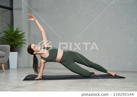 Graceful fit female in sportswear doing side plank exercise, while training at home. Front view of beautiful woman in sports bra standing side forearm plank during yoga session. Concept of yoga. 110293144