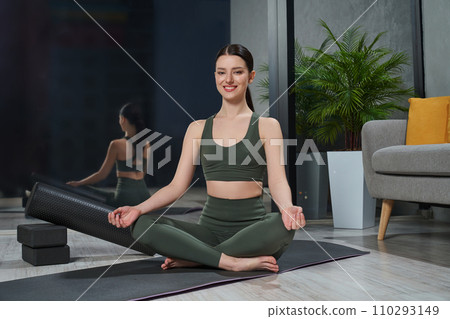 Beautiful smiling girl sitting in Sukhasana position with crossed legs at home. Front view of caucasian lady relaxing in easy yoga pose with mudra hands in modern bedroom on floor. Meditation concept. Beautiful smiling girl sitting in Sukhasana position with crossed legs at home. Front view of caucasian lady relaxing in easy yoga pose with mudra hands in modern bedroom on floor. Meditation concept. 110293149
