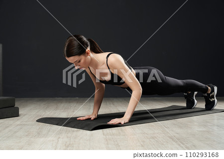 Determined girl in gym clothes getting ready to stay in chaturanga yoga pose during yoga class. Side view of pretty lady with ponytail exercising,in front of black wall indoors. Concept of yoga.  110293168