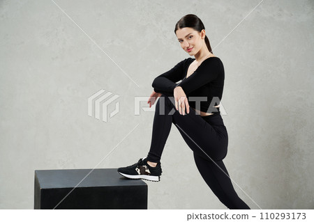 Pretty sporty woman stepping on black cube for fitness and smiling. Side view of fit dark-haired girl with ponytail posing with crossed hands while exercising in studio. Concept of sporty lifestyle. Pretty sporty woman stepping on black cube for fitness and smiling. Side view of fit dark-haired girl with ponytail posing with crossed hands while exercising in studio. Concept of sporty lifestyle. 110293173