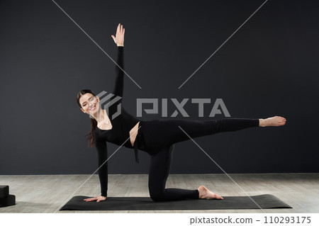Slender brunette girl practicing pose of yoga, stretching on floor, in front of dark wall background. Front view of attractive woman in activewear exercising alone, looking camera. Concept of yoga. 110293175