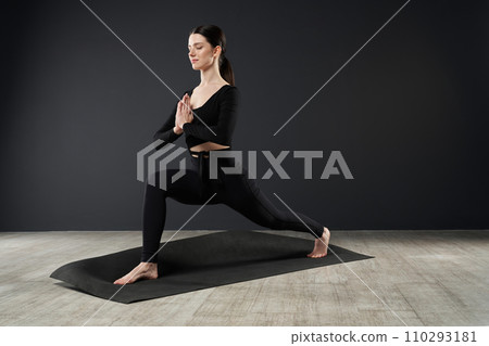 Focused caucasian female lunging with clasped hands on yoga mat indoors. Side view of sporty brunette with folded palms, doing Crescent Lunge pose, during yoga session at home. Concept of yoga. Focused caucasian female lunging with clasped hands on yoga mat indoors. Side view of sporty brunette with folded palms, doing Crescent Lunge pose, during yoga session at home. Concept of yoga. 110293181