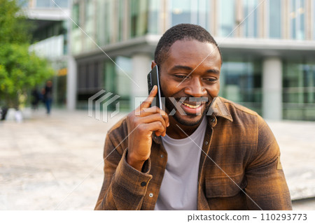 African american man talking on smartphone on street in city outdoor. Man with cell phone chatting with friends. Smiling person making answering call by cellphone. Guy having conversation by mobile African american man talking on smartphone on street in city outdoor. Man with cell phone chatting with friends. Smiling person making answering call by cellphone. Guy having conversation by mobile 110293773