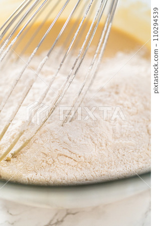 Mixing wet and dry ingredients with a hand whisk in a glass mixing bowl to bake lemon poppy seed muffins. Mixing wet and dry ingredients with a hand whisk in a glass mixing bowl to bake lemon poppy seed muffins. 110294539