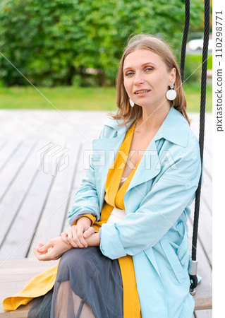 Happy woman swinging on swing in city park, wearing yellow dress and blue coat 110298771