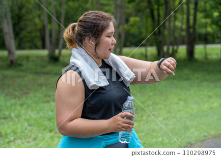 Young overweight woman checking the result of her morning run on her smart watch while standing at running track of a local park Young overweight woman checking the result of her morning run on her smart watch while standing at running track of a local park 110298975