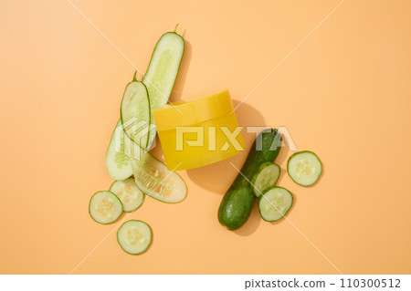 Top view of an unlabeled yellow cosmetic jar and thin cucumber slices on a pastel background. Minimalist cosmetic mockup for advertising. Empty label for branding. 110300512