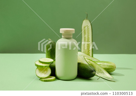 On a green background, an unlabeled cosmetic bottle and fresh cucumbers are displayed. Ideal space for advertising beauty products with natural extracts. 110300513
