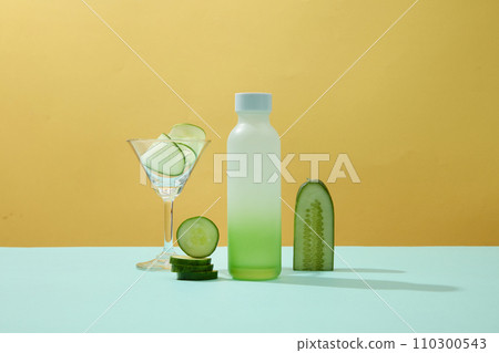 A blank plastic bottle, cocktail glass, and fresh cucumber slices adorn a pastel background-perfect mockups for imaginative advertising. Front view. A blank plastic bottle, cocktail glass, and fresh cucumber slices adorn a pastel background-perfect mockups for imaginative advertising. Front view. 110300543