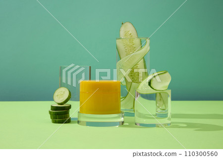 Front view of an unlabeled yellow cosmetic jar placed on a glass platform, surrounded by glasses with thin slices of cucumber on a blue background. 110300560
