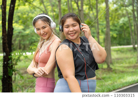 Two happy Asian female friends, one over size and one lean, standing with their back together listening to music from their headphones at a local park Two happy Asian female friends, one over size and one lean, standing with their back together listening to music from their headphones at a local park 110303315