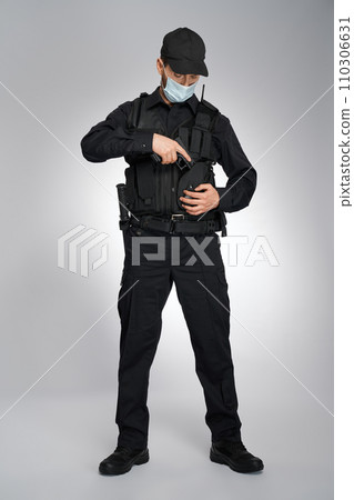 Confident policeman in uniform hiding handgun in holster indoors. Serious police officer in protective mask looking down, while removing gun, on gray studio background. Concept of work, quarantine. Confident policeman in uniform hiding handgun in holster indoors. Serious police officer in protective mask looking down, while removing gun, on gray studio background. Concept of work, quarantine. 110306631