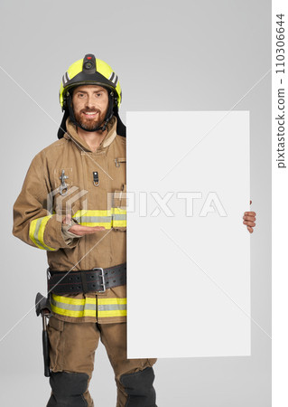 Happy firefighter in uniform standing with blank cardboard indoors. Front view of smiling caucasian fireman demonstrating empty banner, isolated on gray background. Concept of announcement. Happy firefighter in uniform standing with blank cardboard indoors. Front view of smiling caucasian fireman demonstrating empty banner, isolated on gray background. Concept of announcement. 110306644