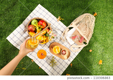 Picnic basket with fruits, flowers, lipsticks and book on the white checkered mat in the green park background. Hand of woman is holding a cool juice cups 110306863