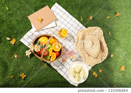 Top view of wicker picnic basket with fruits, hat and books on white checkered tablecloth on green grass outside in summer park. Advertising photo, summer day concept 110306864