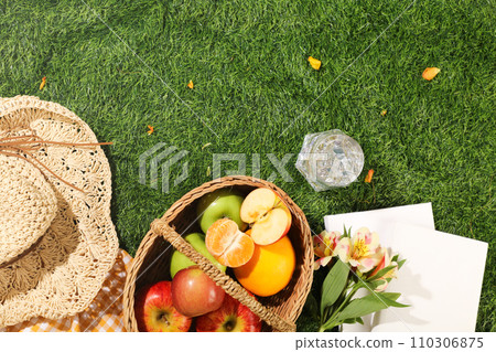Delicious picnic spread with basket of fruits, hat, cup of cool water and white notebooks on green grass background. Top view, holiday summer concept. Space for design Delicious picnic spread with basket of fruits, hat, cup of cool water and white notebooks on green grass background. Top view, holiday summer concept. Space for design 110306875