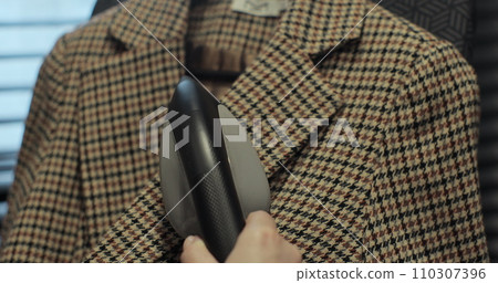 Woman steams plaid jacket with electric steamer at home. Dry cleaning clothes. Household and home equipment for housework. Woman steams plaid jacket with electric steamer at home. Dry cleaning clothes. Household and home equipment for housework. 110307396