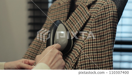 Woman steams plaid jacket with electric steamer at home. Dry cleaning clothes. Household and home equipment for housework. Woman steams plaid jacket with electric steamer at home. Dry cleaning clothes. Household and home equipment for housework. 110307400