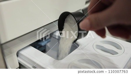 Woman pours washing powder into the washing machine compartment. Female with measuring spoon, fills the washing machine with powder. 110307763