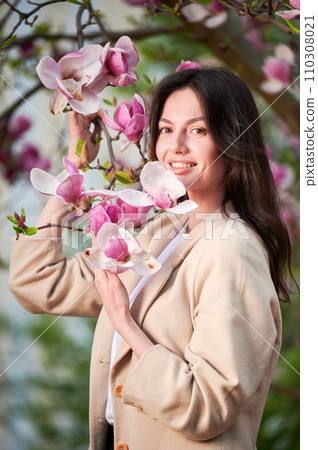 Woman allergic enjoying after treatment from seasonal allergy at spring. Portrait of happy young woman smiling in front of blooming magnolia tree at springtime. Spring allergy concept. Woman allergic enjoying after treatment from seasonal allergy at spring. Portrait of happy young woman smiling in front of blooming magnolia tree at springtime. Spring allergy concept. 110308021