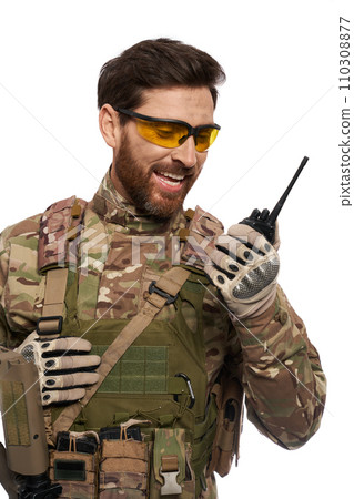 Excited soldier in armor vest, talking on walkie talkie in studio. Portrait of smiling infantryman in uniform and glasses, using portable radio, reporting, isolated on white. Army, connection concept. 110308877
