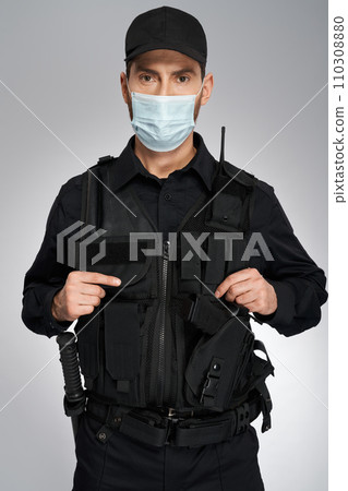 Young caucasian policeman wearing protective mask looking at camera. Portrait of confident police officer in bulletproof vest posing, isolated on gray studio background. Concept of work, quarantine. Young caucasian policeman wearing protective mask looking at camera. Portrait of confident police officer in bulletproof vest posing, isolated on gray studio background. Concept of work, quarantine. 110308880