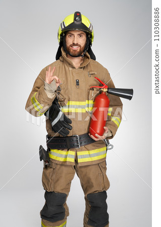 Happy firefighter with extinguisher showing okay sign indoors. Front view of bearded fireman in uniform smiling, while gesturing ok, on gray studio background. Firefighting, hand gesture concept. Happy firefighter with extinguisher showing okay sign indoors. Front view of bearded fireman in uniform smiling, while gesturing ok, on gray studio background. Firefighting, hand gesture concept. 110308886