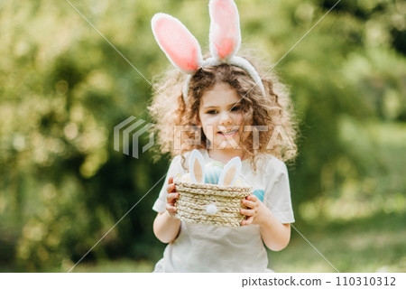 Easter egg hunt. Girl child Wearing Bunny Ears Running To Pick Up Egg In Garden. Easter tradition. Baby with basket full of colorful eggs. Easter egg hunt. Girl child Wearing Bunny Ears Running To Pick Up Egg In Garden. Easter tradition. Baby with basket full of colorful eggs. 110310312