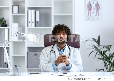 Webcam view, young doctor with headset phone using laptop for video call, doctor cheerfully and friendly consulting patients, smiling and looking at camera, working inside clinic medical room. 110314422