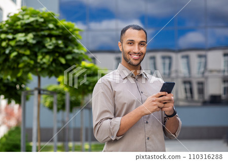 Portrait of young businessman, man smiling looking at camera holding phone, worker using app on smartphone while walking in city close up outside office building. 110316288