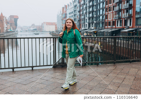 Attractive young female tourist is exploring new city. Redhead girl with backpack holding a paper map on city street in Gdansk. Traveling Europe in autumn. Famous Zuraw crane, Motlawa river 110316640