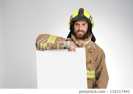 Smiling firefighter in protective helmet holding blank banner in studio. Portrait of bearded man in uniform demonstrating big white sheet with copy space, isolated on gray. Work, copy place concept. 110317491