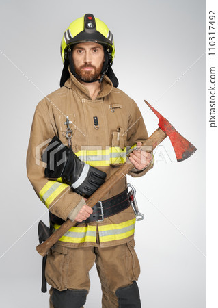 Bearded caucasian firefighter in full gear holding red axe for fire fighting in studio. Front view of handsome fireman with hatchet looking at camera, on gray background. Concept of work, fire tools. 110317492