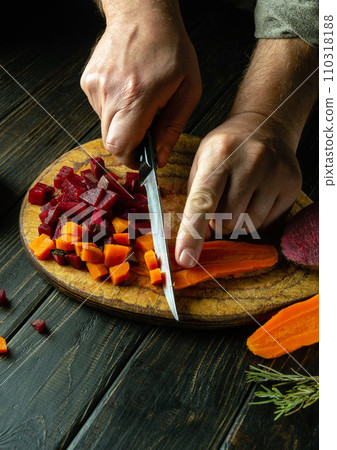The chef uses a knife to cut boiled carrots to prepare a vegetable dish on the kitchen table. Venaigret cooking concept on dark background The chef uses a knife to cut boiled carrots to prepare a vegetable dish on the kitchen table. Venaigret cooking concept on dark background 110318188