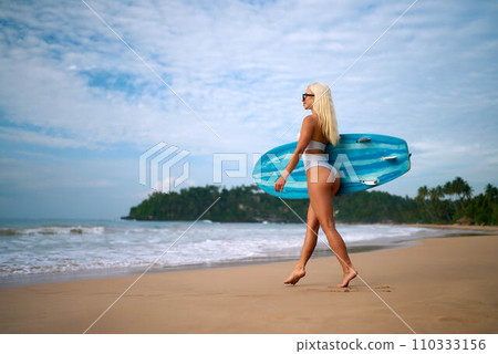Blonde woman with sunglasses walks on sandy beach carrying blue surfboard, ready to surf waves by tropical coast. Female surfer enjoys summer day at sea shore preparing for water sports. Blonde woman with sunglasses walks on sandy beach carrying blue surfboard, ready to surf waves by tropical coast. Female surfer enjoys summer day at sea shore preparing for water sports. 110333156