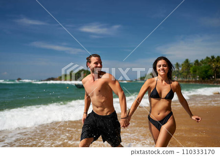 Fit couple runs on tropical beach, playful man and woman exercise near ocean. Athletic pair in swimwear enjoys summer vacation, jogging along shoreline, healthy lifestyle, beach workout, sunny day. 110333170