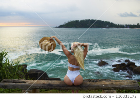 Woman in bikini with a beautiful physique sitting on beach looking at ocean view on sunset in swimwear. Slim figure girl enjoys sea vacation with straw hat in hands. Pretty slender fit blonde female. 110333172