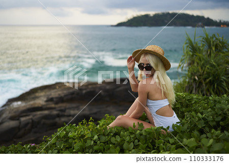 Joyful woman enjoying ocean vistas, adjusts straw hat. Wearing white swimwear, she sits by sea, lush foliage foreground, embodying leisure, tropical travel, and the concept of wanderlust. 110333176