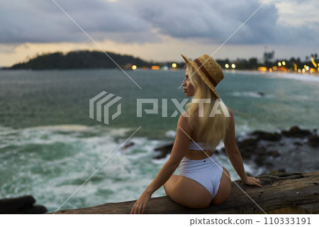 Blonde woman in white bikini and straw hat sits on seaside cliff at dusk, gazing over turbulent waves, embodying solo travel and serene contemplation by the ocean Blonde woman in white bikini and straw hat sits on seaside cliff at dusk, gazing over turbulent waves, embodying solo travel and serene contemplation by the ocean 110333191