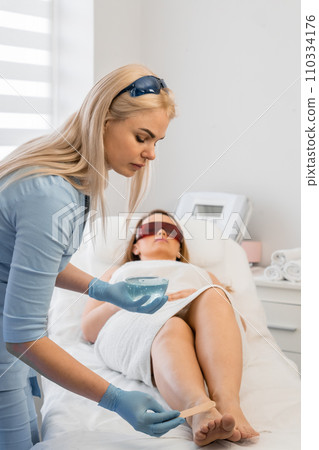 The beautician applies a special substance to the woman's skin before the rocedura. In a beauty salon, a beautician performs photoepilation woman's skin using laser technology for impeccable result. 110334176