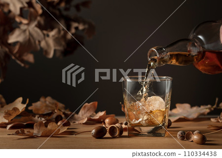 Whiskey with ice on a wooden table with dried-up oak leaves. 110334438