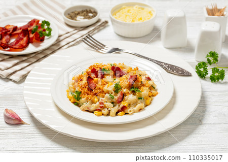 portion of mac and cheese with corn, bacon topped with panko breadcrumbs on white plate on white wooden table  110335017