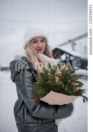 Portrait of young blond woman in beige coat walking in the city with a bouquet of fresh fir branches Portrait of young blond woman in beige coat walking in the city with a bouquet of fresh fir branches 110336831