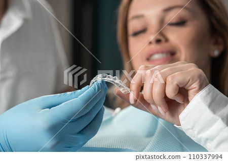 Attentive dentist demonstrating aligner aligner in hands, showing female patient in dental office, preparing for work and striving to make the procedure as comfortable as possible for her patient. Attentive dentist demonstrating aligner aligner in hands, showing female patient in dental office, preparing for work and striving to make the procedure as comfortable as possible for her patient. 110337994