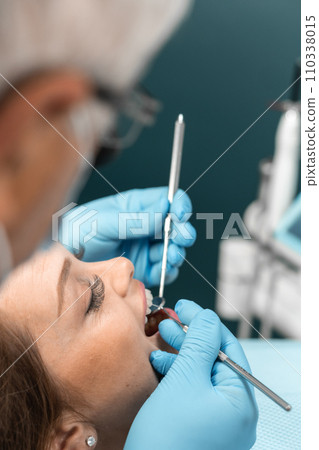 A patient sits in a dental chair with a smile, expressing her impression of refreshment and lightness after a visit to the dentist. The doctor, holding instruments in his hands, is ready for further A patient sits in a dental chair with a smile, expressing her impression of refreshment and lightness after a visit to the dentist. The doctor, holding instruments in his hands, is ready for further 110338015